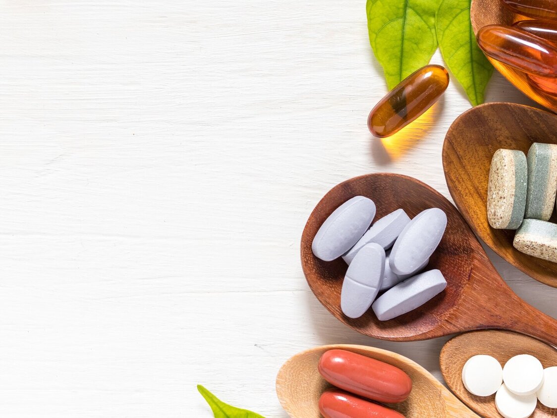 Auf einem weißen Untergrund liegen fünf Holzlöffel mit verschiedenen Arten von Tabletten und Kapseln. 