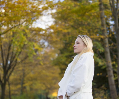 Eine blonde Frau mit weißem Mantel läuft durch einen herbstlichen Wald und atmet tief ein.