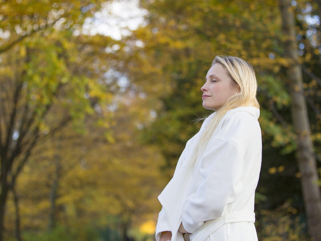 Eine blonde Frau mit weißem Mantel läuft durch einen herbstlichen Wald und atmet tief ein.