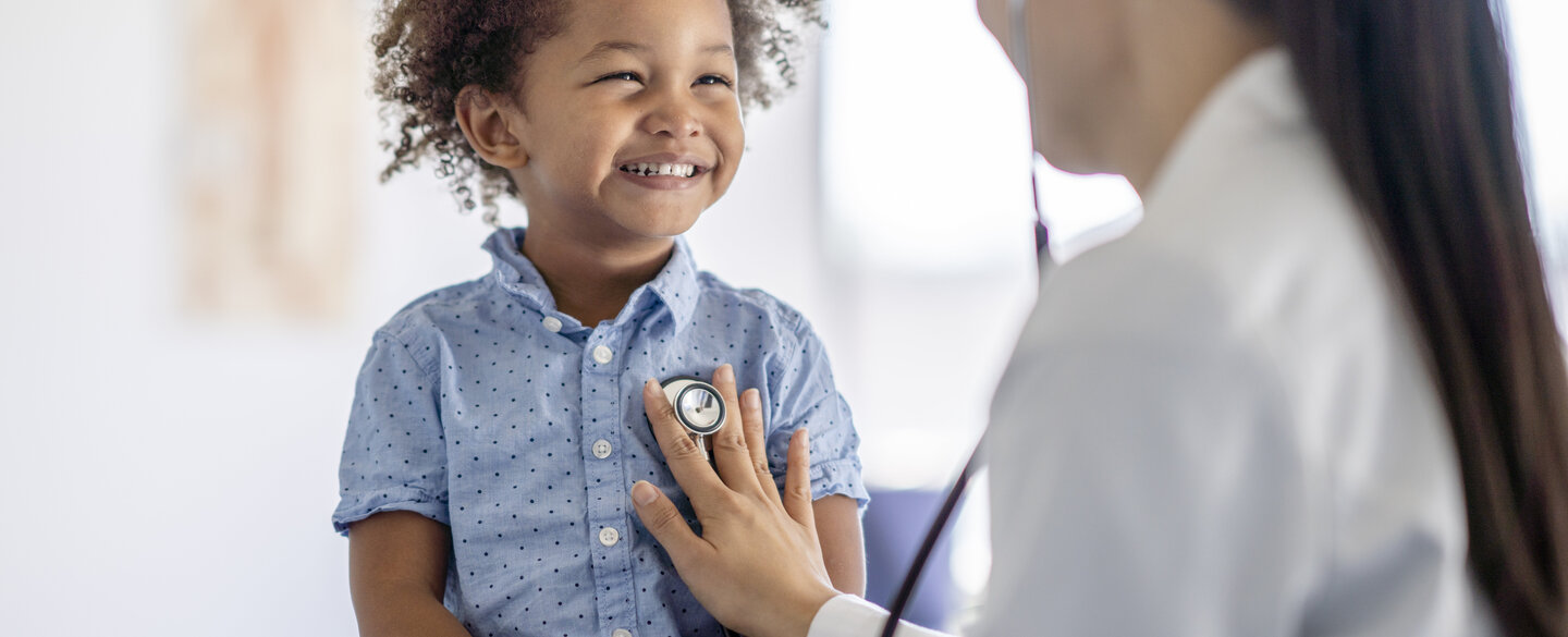 Eine Ärztin asiatischer Abstammung hört mit ihrem Stethoskop auf das Herz ihrer Patienten. Der kleine Junge sitzt oben auf dem Untersuchungstisch und lächelt seiner Ärztin zu, während sie das Instrument an seine Brust hält.