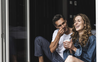 Ein Mann und eine Frau sitzend lachend an der Terasse. Sie trinkt Kaffee und er hat einen Löffel am Mund.