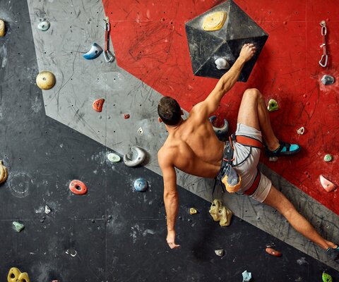 Ein Mann hängt an einer Kletterwand und hält sich mit einer Hand fest.