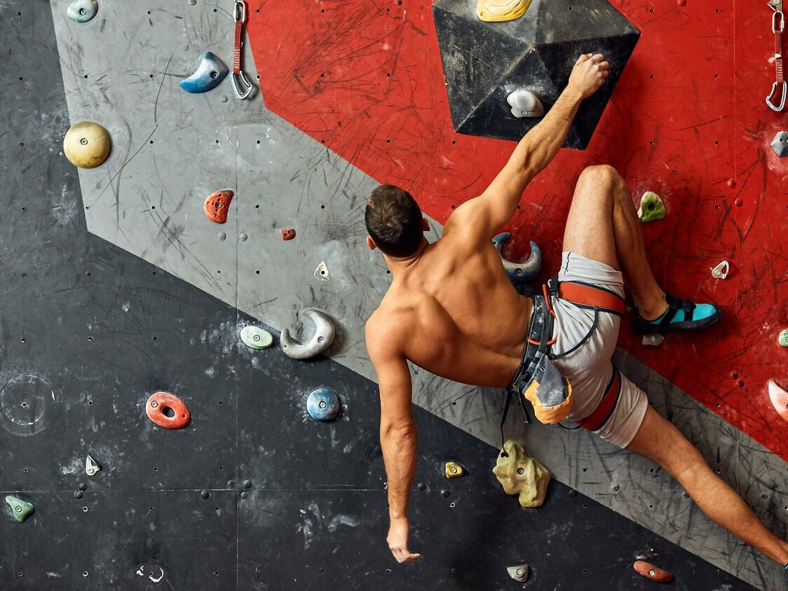 Ein Mann hängt an einer Kletterwand und hält sich mit einer Hand fest.