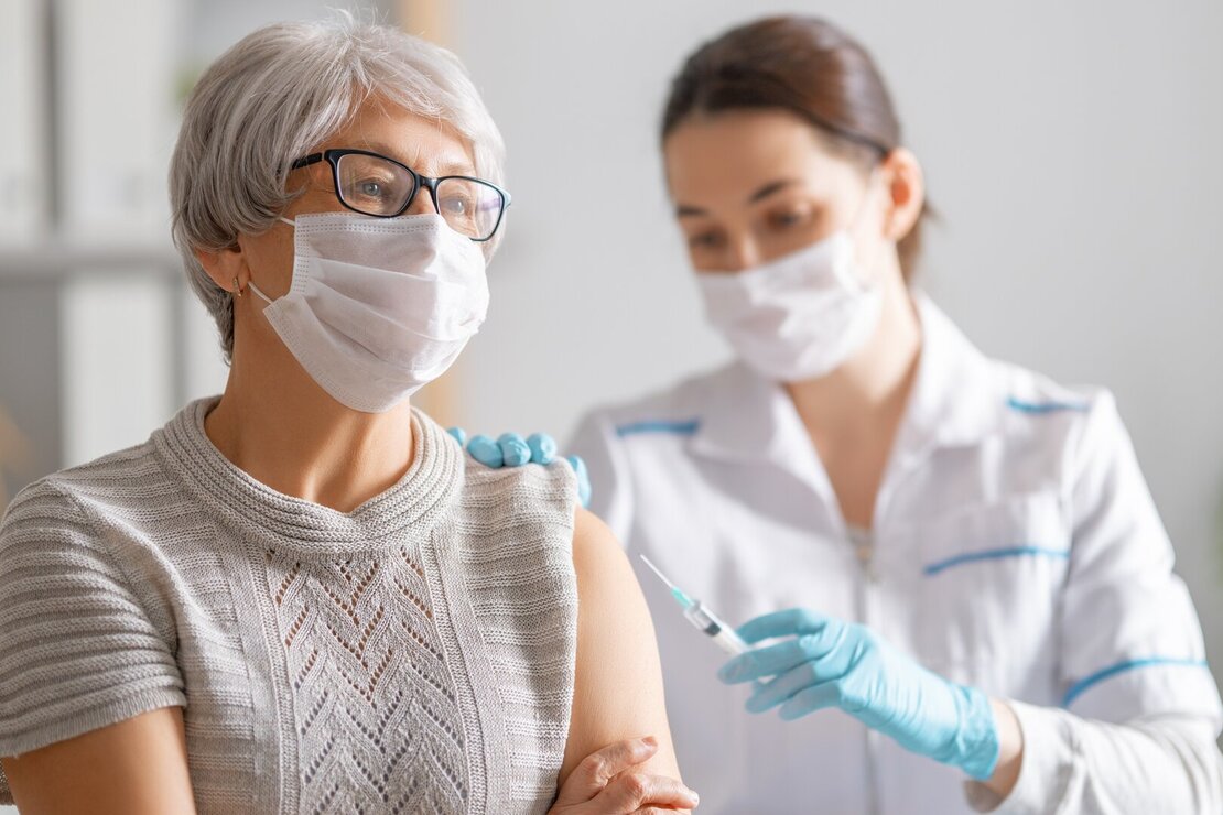 Ältere Damen mit Mundschutz bekommt von einer Ärztin mit Mundschutz eine Spritze in den Oberarm.