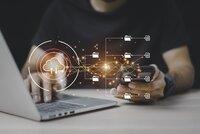 Jemand tippt auf einem Laptop und hat dabei ein Handy in der anderen Hand. Im Vordergrund werden virtuelle Symbole angezeigt.