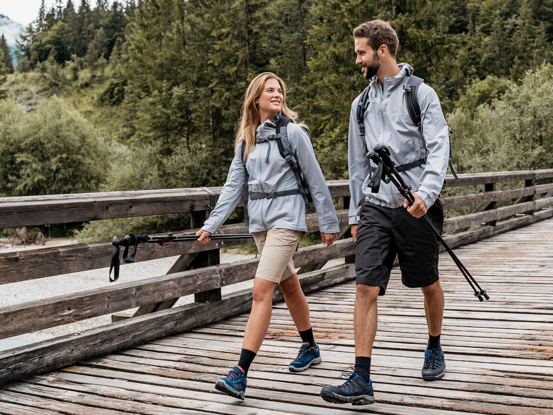 Ein Paar bei einer Wanderung über einen Holzsteg. Sie haben Stöcke in den Händen und einen Rucksack auf dem Rücken. Im Hintergrund ist ein Wald.