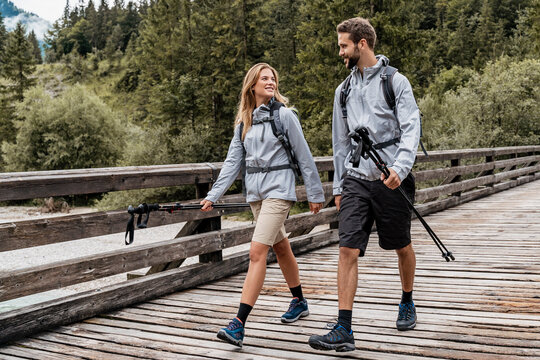 Ein Paar bei einer Wanderung über einen Holzsteg. Sie haben Stöcke in den Händen und einen Rucksack auf dem Rücken. Im Hintergrund ist ein Wald.