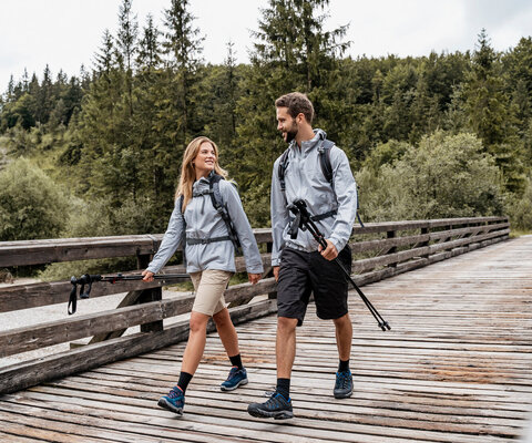 Ein Paar bei einer Wanderung über einen Holzsteg. Sie haben Stöcke in den Händen und einen Rucksack auf dem Rücken. Im Hintergrund ist ein Wald.