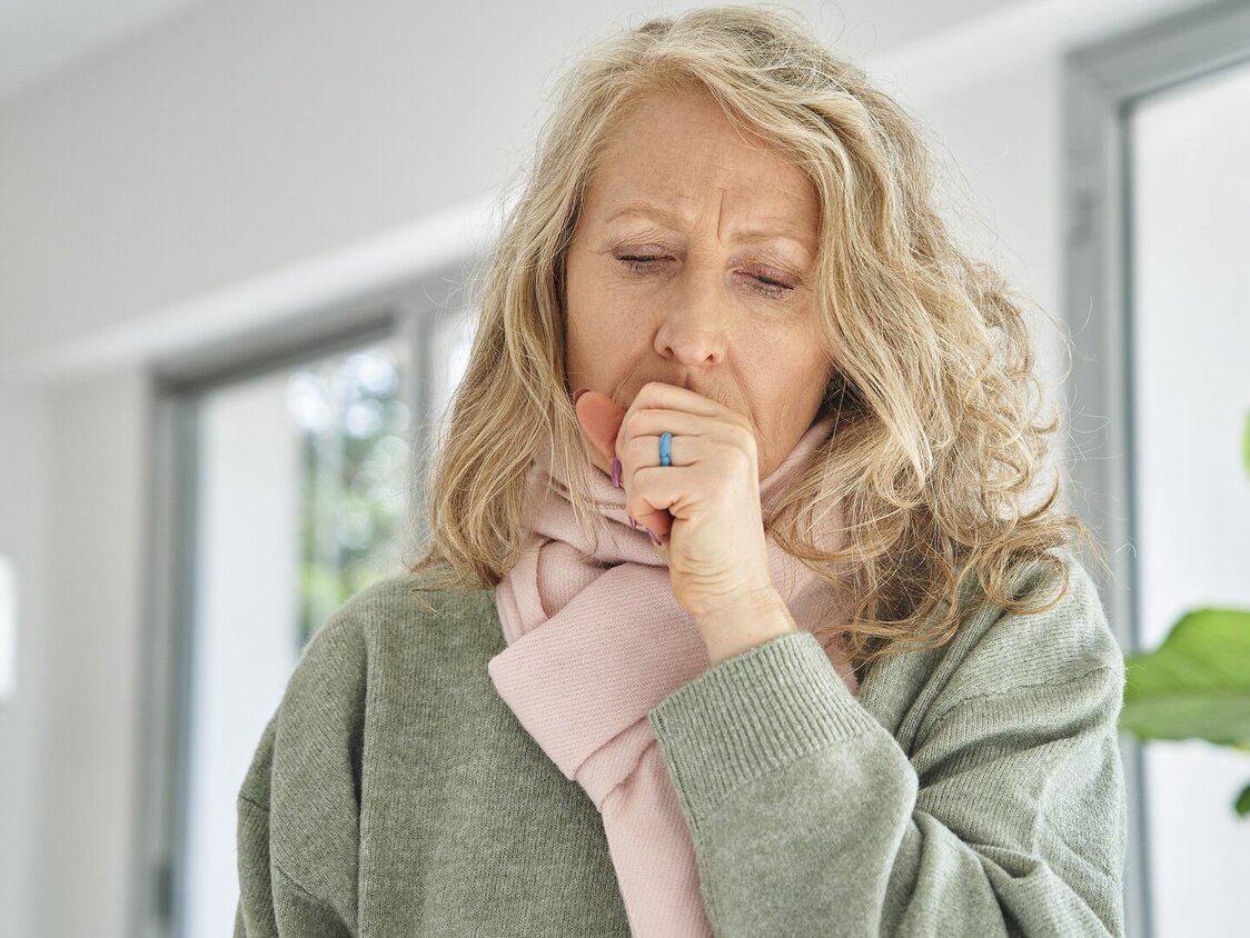 Frau mit Schal hustet in ihre Faust während sie in einem hellen Raum steht.