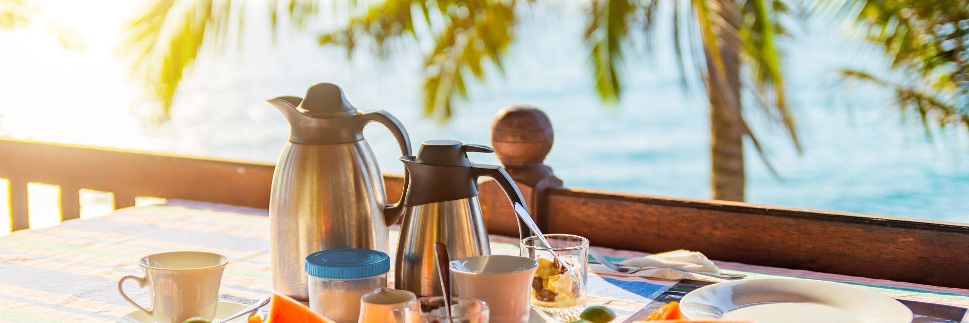 Auf einem Tisch stehen eine Kaffeekanne, eine Teekanne, Tassen und Teller mit Obst. Im Hintergrund sind das Meer und eine Palme.