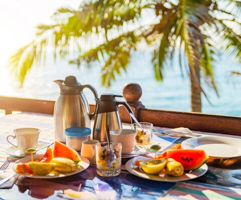 Auf einem Tisch stehen eine Kaffeekanne, eine Teekanne, Tassen und Teller mit Obst. Im Hintergrund sind das Meer und eine Palme.