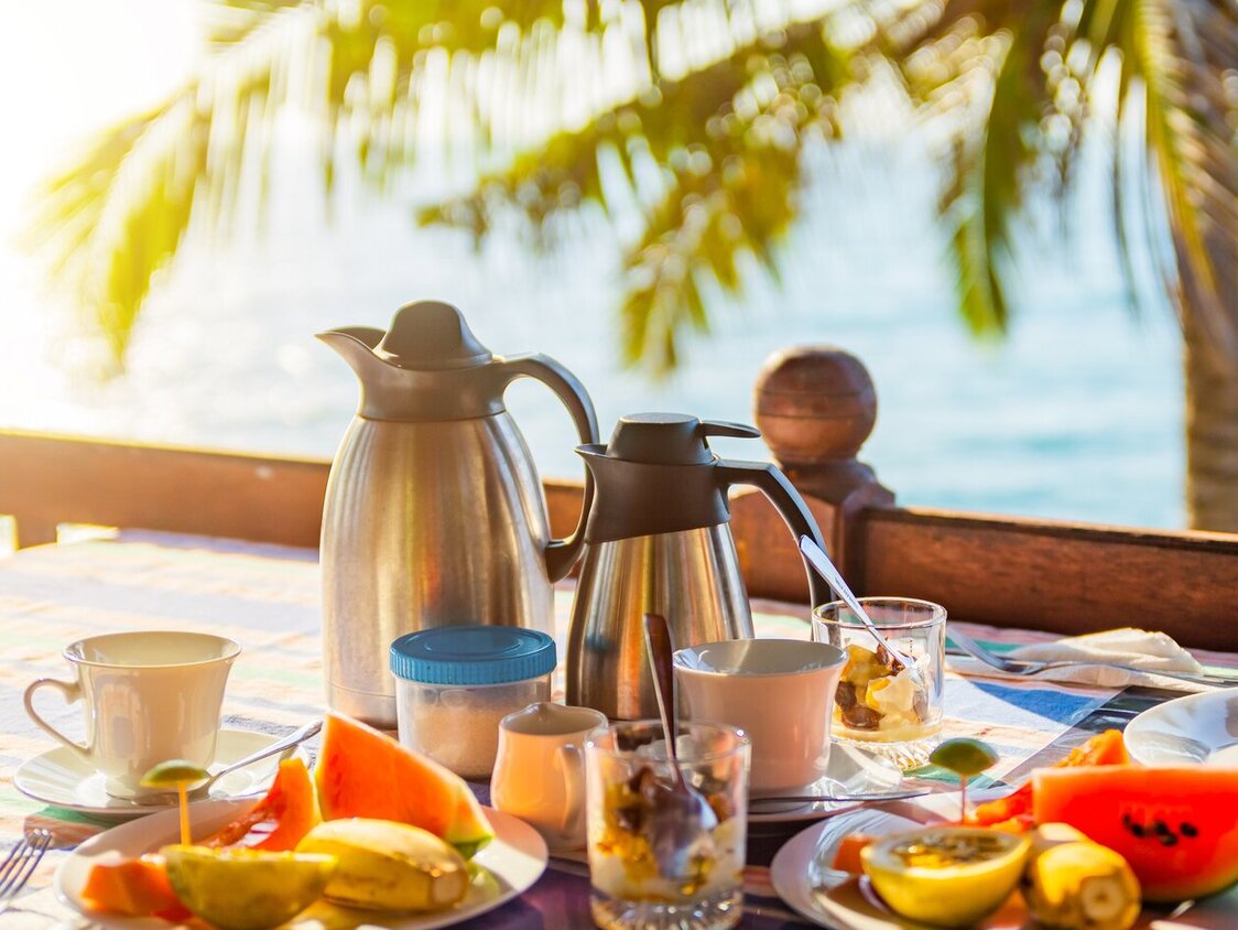 Auf einem Tisch stehen eine Kaffeekanne, eine Teekanne, Tassen und Teller mit Obst. Im Hintergrund sind das Meer und eine Palme.
