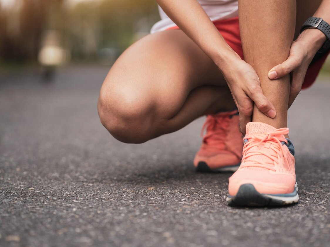 Eine Frau in Joggingkleidung kniet und hält sich das Fußgelenk.