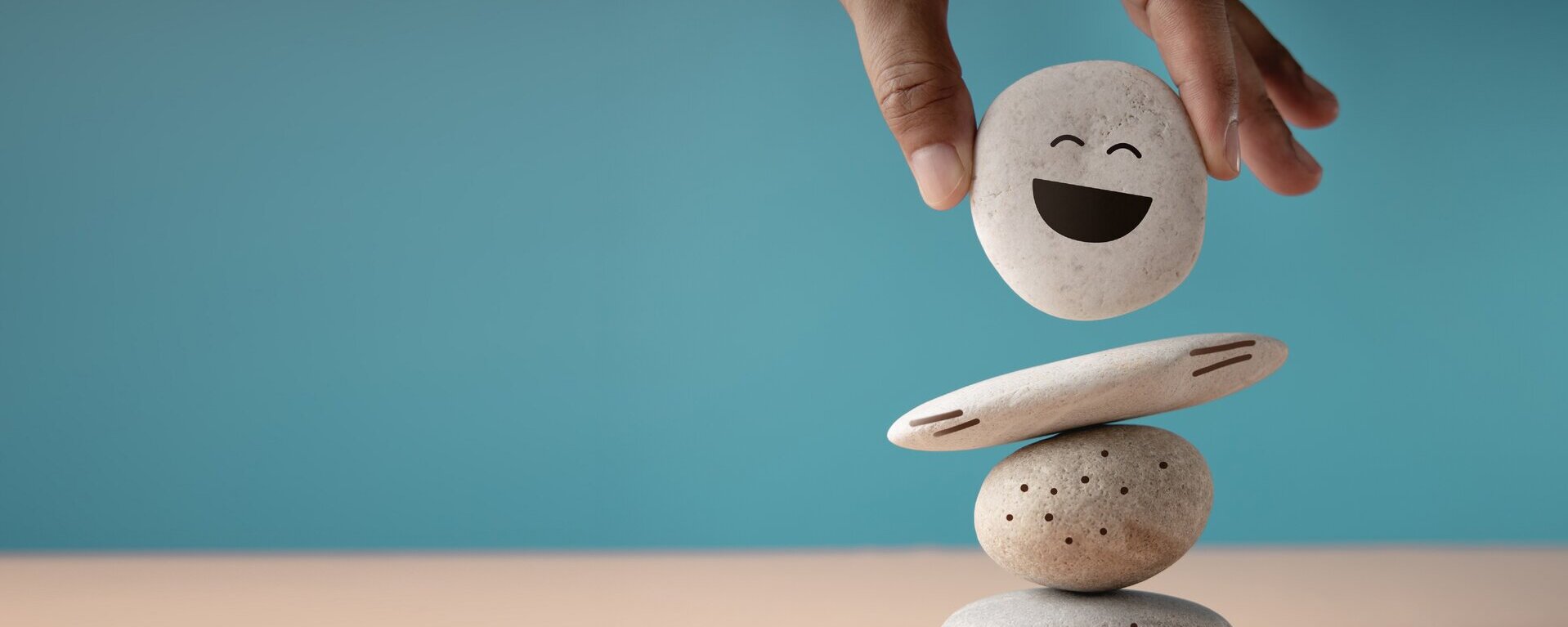 Ein Steinmännchen aus vier Steinen mit einem Gesicht darauf gemalt. Eine Hand setzt den letzten Stein darauf.