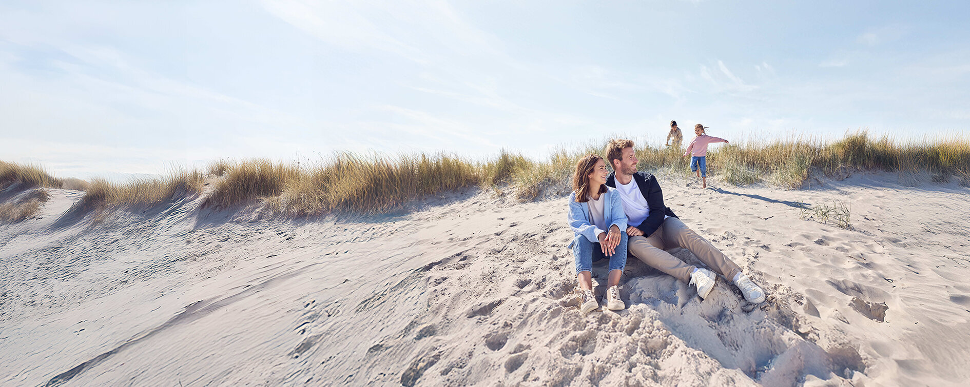 Eltern sitzen am Strand und die zwei Kinder spielen n der Düne.