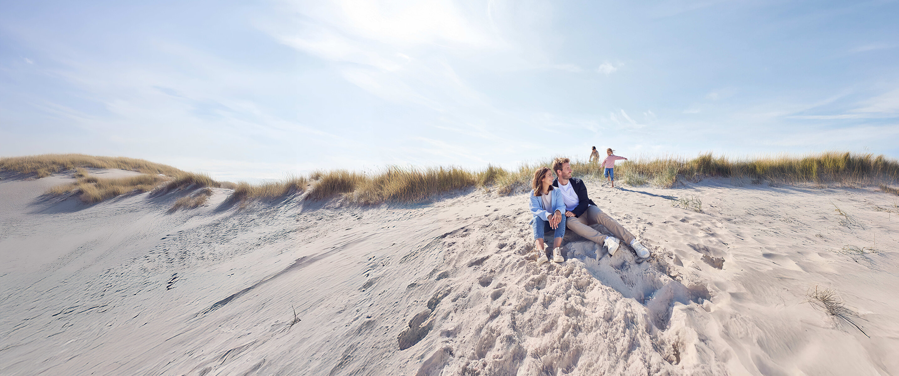 Eltern sitzen am Strand und die zwei Kinder spielen n der Düne.