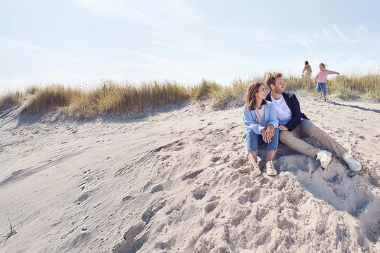 Eltern sitzen am Strand und die zwei Kinder spielen n der Düne.