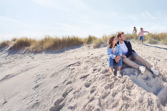 Eltern sitzen am Strand und die zwei Kinder spielen n der Düne.