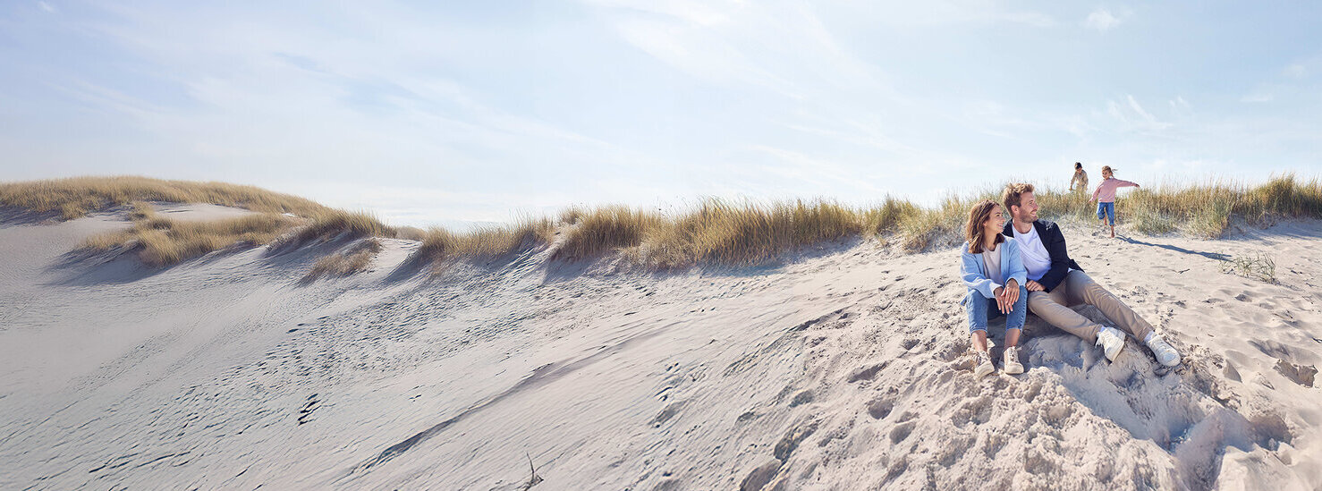 Eltern sitzen am Strand und die zwei Kinder spielen n der Düne.