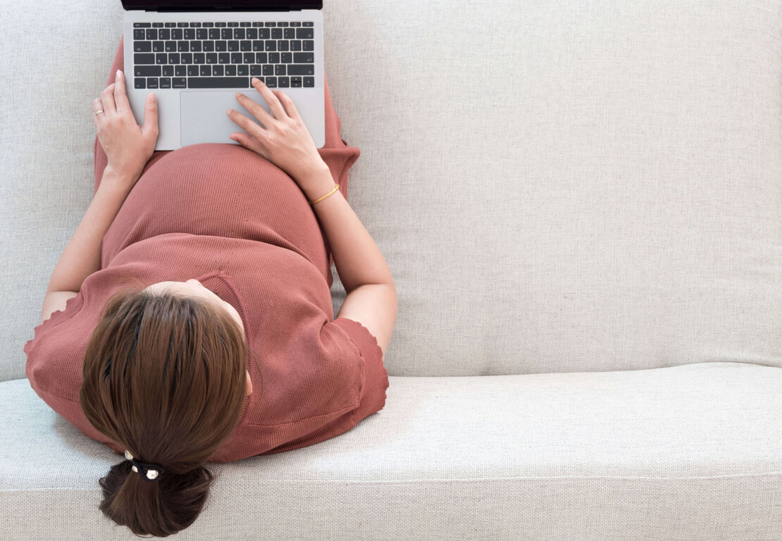  Eine schwangere Frau auf einem Sofa am Laptop sitzend.