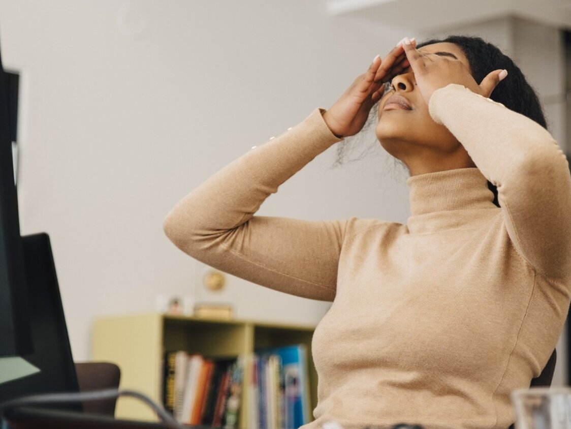 Eine Frau hält sich die Hände über den Kopf. Sie sitzt an ihrem Arbeitsplatz vor einem PC.