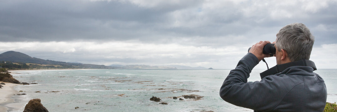 Ein älterer Mann mit grauen Haaren schaut durchs Fernglas über die Meeresbucht. 
