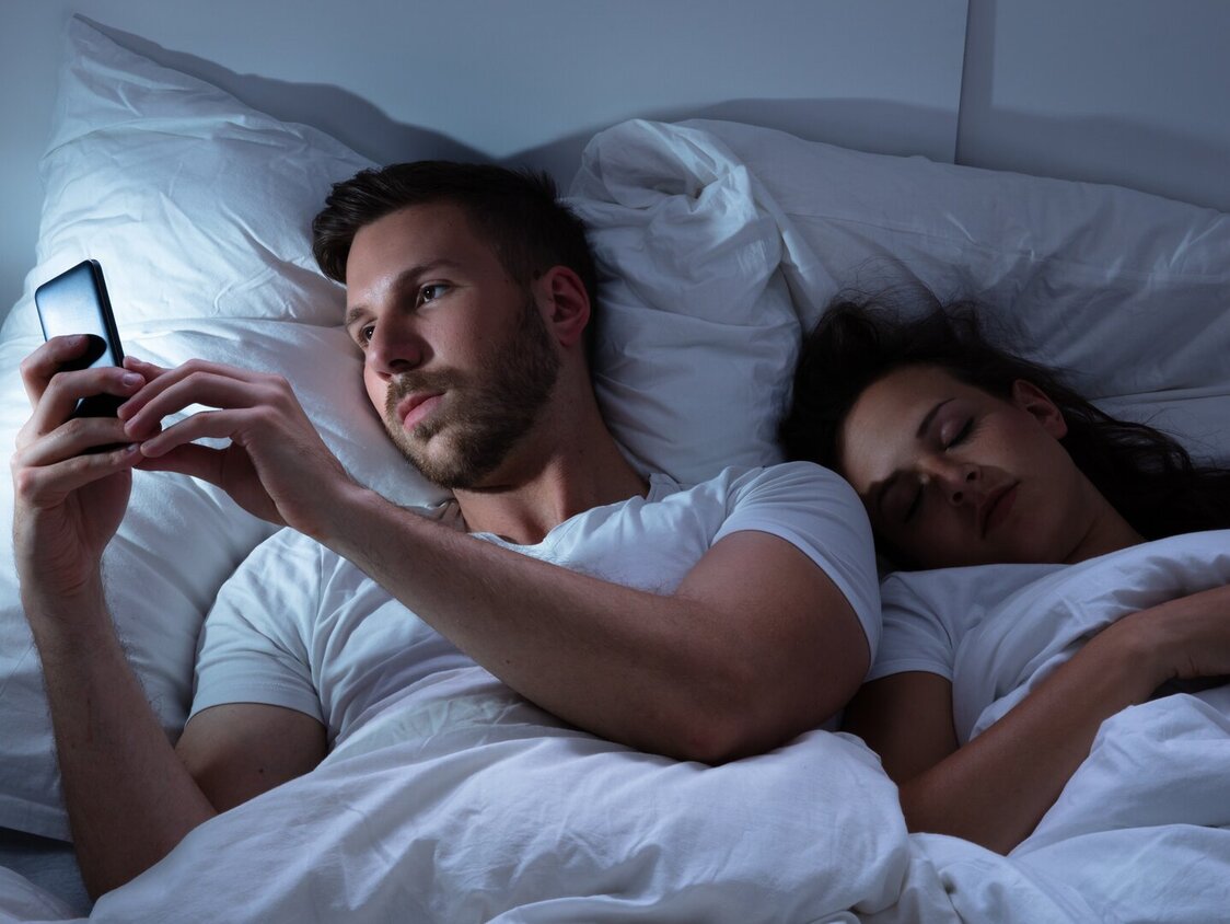 Ein Mann liegt im Bett neben einer schlafenden Frau und tippt auf seinem Handy.