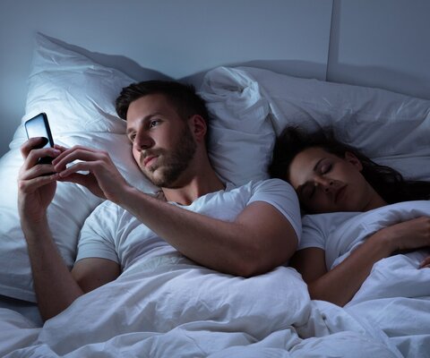 Ein Mann liegt im Bett neben einer schlafenden Frau und tippt auf seinem Handy.