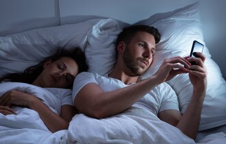 Ein Mann liegt im Bett neben einer schlafenden Frau und tippt auf seinem Handy.