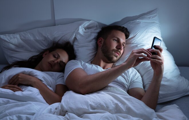 Ein Mann liegt im Bett neben einer schlafenden Frau und tippt auf seinem Handy.