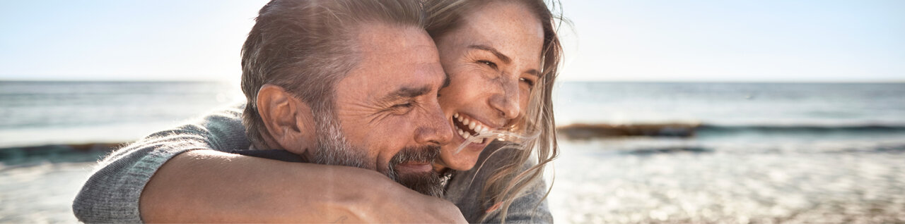 Eine Frau und ein Mann umarmen sich lachend am Strand.
