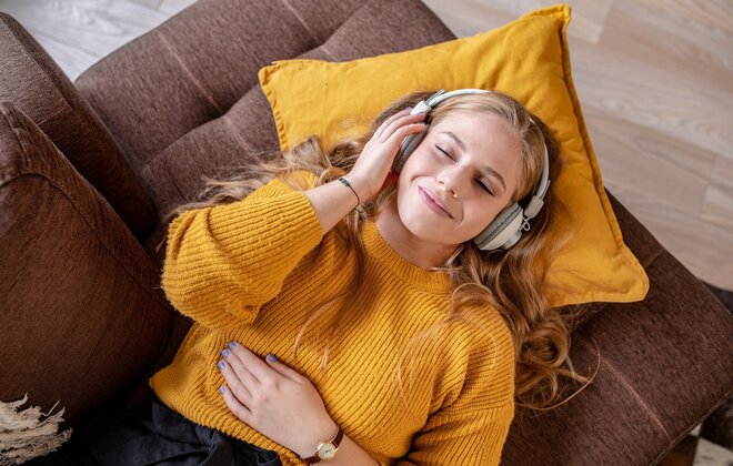 Eine Frau liegt mit geschlossenen Augen auf dem Sofa und hat Kopfhörer auf.