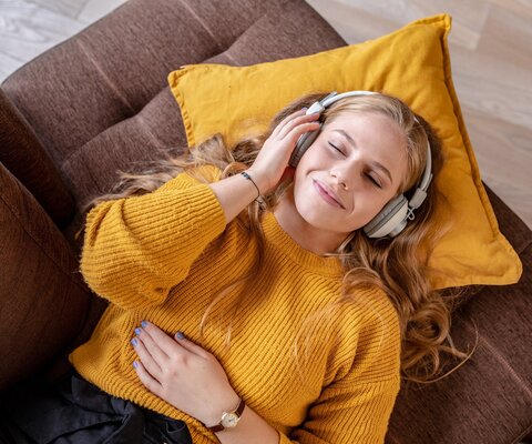 Eine Frau liegt mit geschlossenen Augen auf dem Sofa und hat Kopfhörer auf.