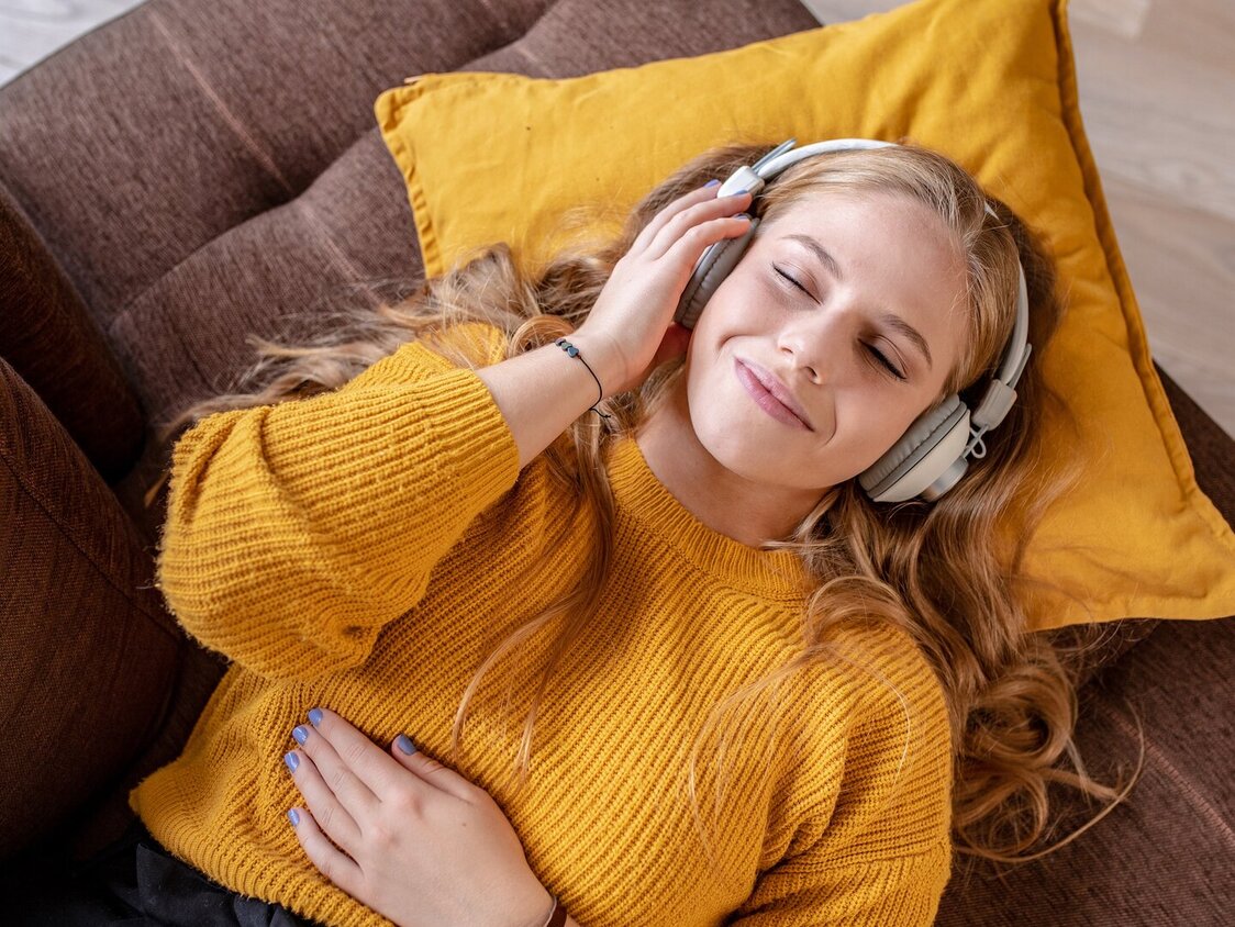 Eine Frau liegt mit geschlossenen Augen auf dem Sofa und hat Kopfhörer auf.