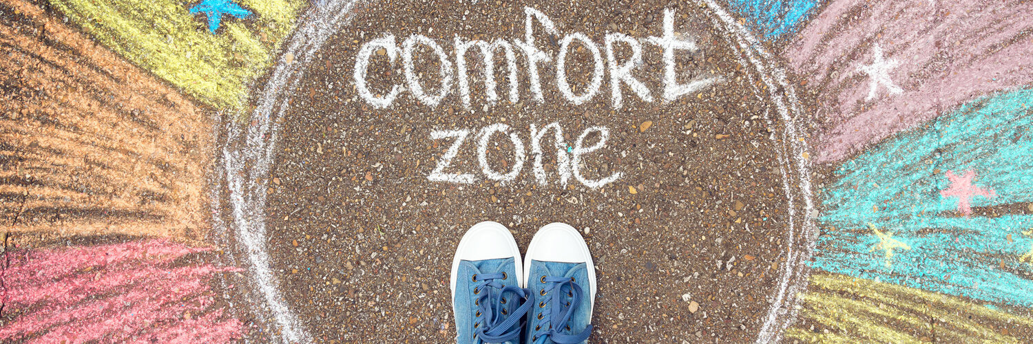 Eine Person steht in einem Kreis, welcher durch Kreide auf den Boden gezeichnet wurde. Darin steht der Begriff comfort zone. Außerhalb des Kreises sind bunte Kreidefarben und Sterne.