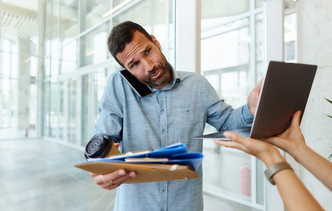 Ein Mann steht in einem Büro: in einer Hand hat er verschiedene Dokumente und einen Kaffee, während er mit der anderen Hand an einem Laptop tippt, der ihm hingehalten wird. Zwischen Schulter und Ohr hat er außerdem ein Smartphone geklemmt.