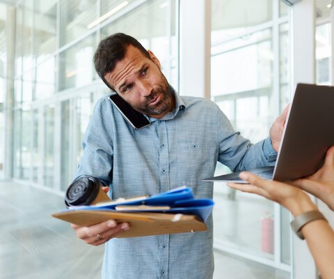 Ein Mann steht in einem Büro: in einer Hand hat er verschiedene Dokumente und einen Kaffee, während er mit der anderen Hand an einem Laptop tippt, der ihm hingehalten wird. Zwischen Schulter und Ohr hat er außerdem ein Smartphone geklemmt.