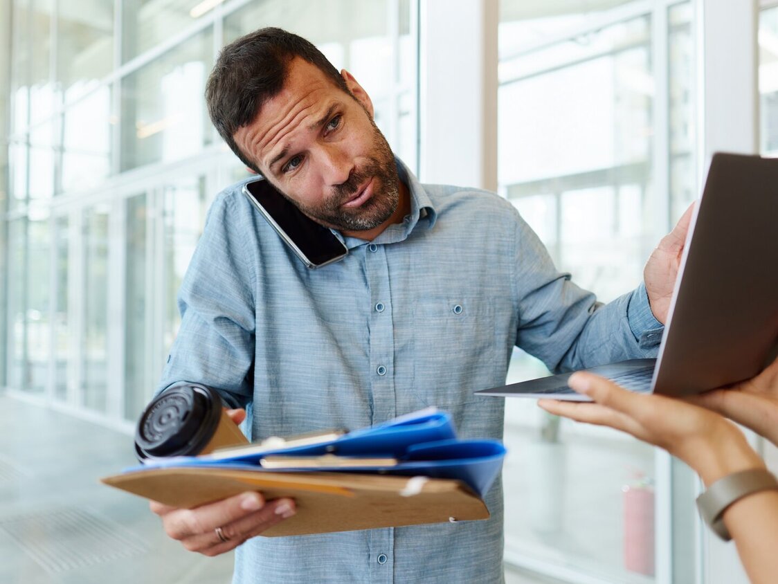 Ein Mann steht in einem Büro: in einer Hand hat er verschiedene Dokumente und einen Kaffee, während er mit der anderen Hand an einem Laptop tippt, der ihm hingehalten wird. Zwischen Schulter und Ohr hat er außerdem ein Smartphone geklemmt.