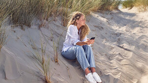 Eine Frau sitzt an einer Düne und guckt auf ihr Handy.