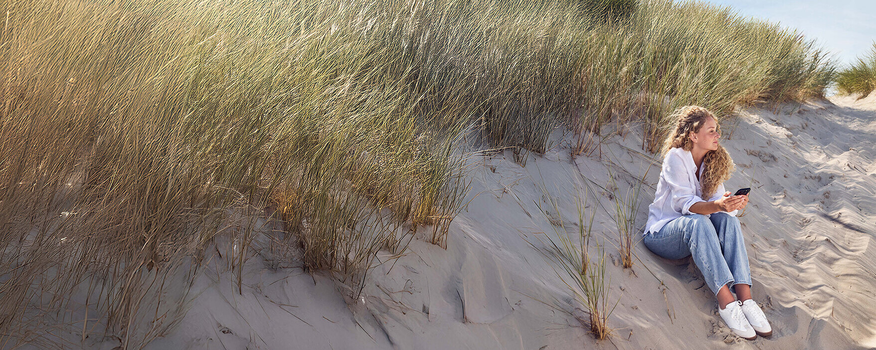 Eine Frau sitzt an einer Düne und guckt auf ihr Handy.