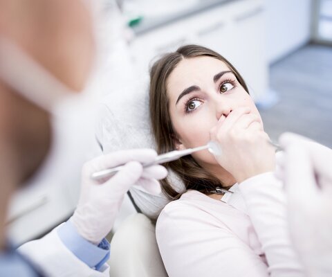 Eine Frau auf dem Behandlungsstuhl und blickt einen Zahnarzt erschrocken an.