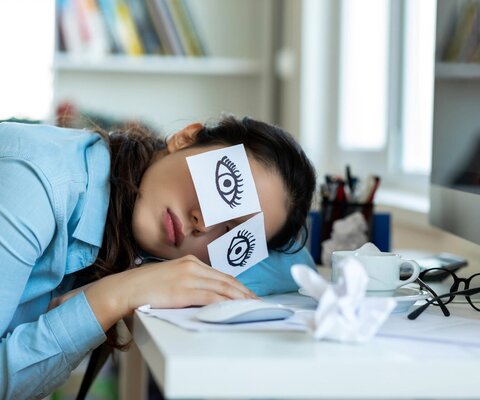 Müde Frau liegt mit Ihrem Kopf auf einem Tisch. Auf Ihren Augen kleben zwei Notizzettel mit Augen drauf gezeichnet.