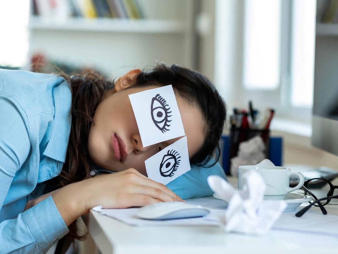 Müde Frau liegt mit Ihrem Kopf auf einem Tisch. Auf Ihren Augen kleben zwei Notizzettel mit Augen drauf gezeichnet.