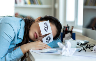 Müde Frau liegt mit Ihrem Kopf auf einem Tisch. Auf Ihren Augen kleben zwei Notizzettel mit Augen drauf gezeichnet.