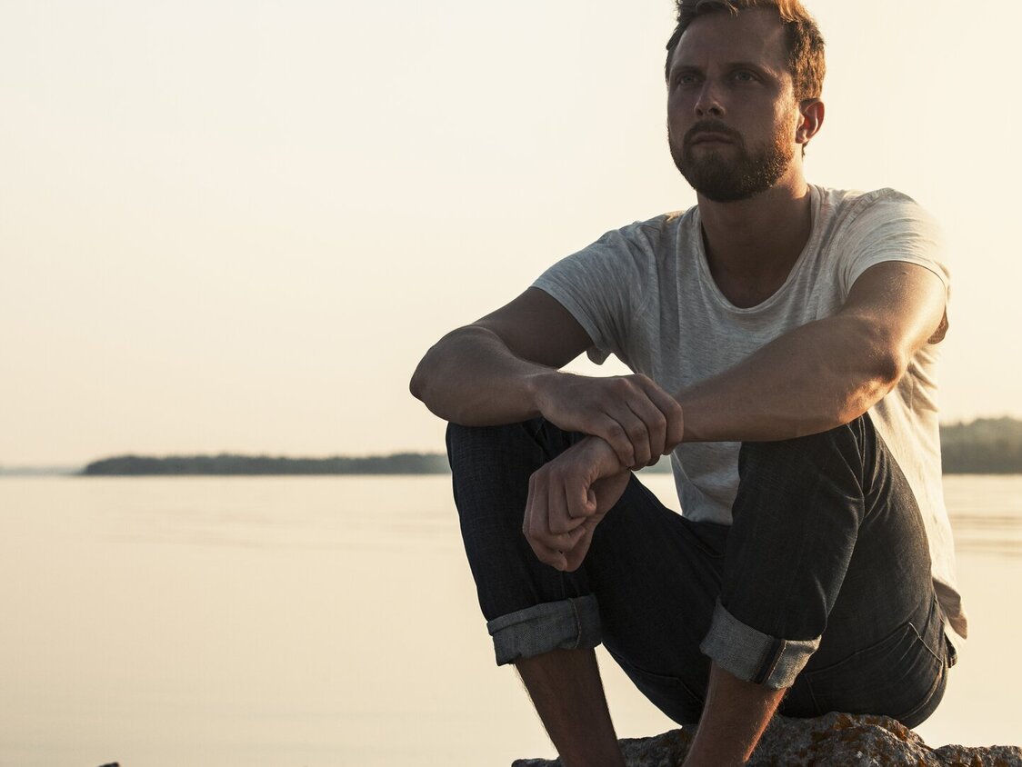 Mann sitzt am Wasser auf Steinen und schaut in die Ferne.