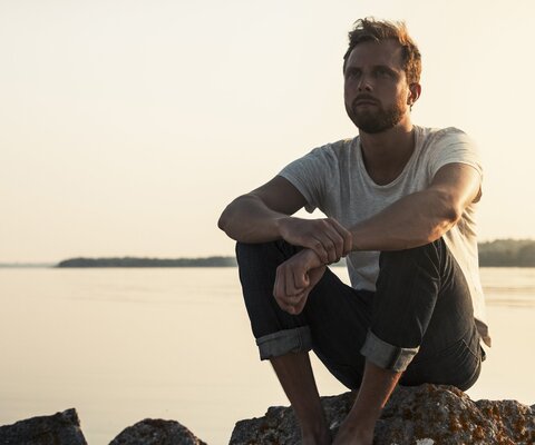 Mann sitzt am Wasser auf Steinen und schaut in die Ferne.