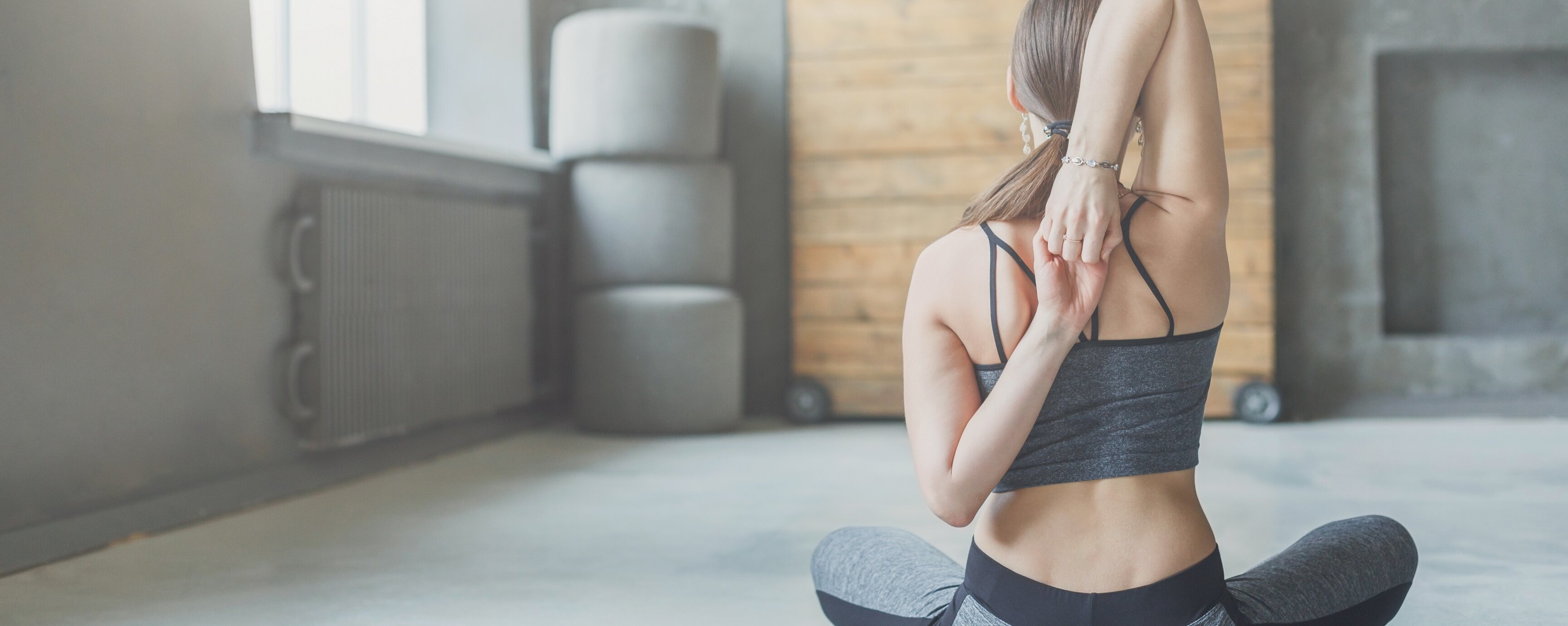 Eine Frau sitzt im Schneidersitz in Sportbekleidung in einem Studio. Man sieht ihren Rücken und sie verschränkt die Arme dahinter.