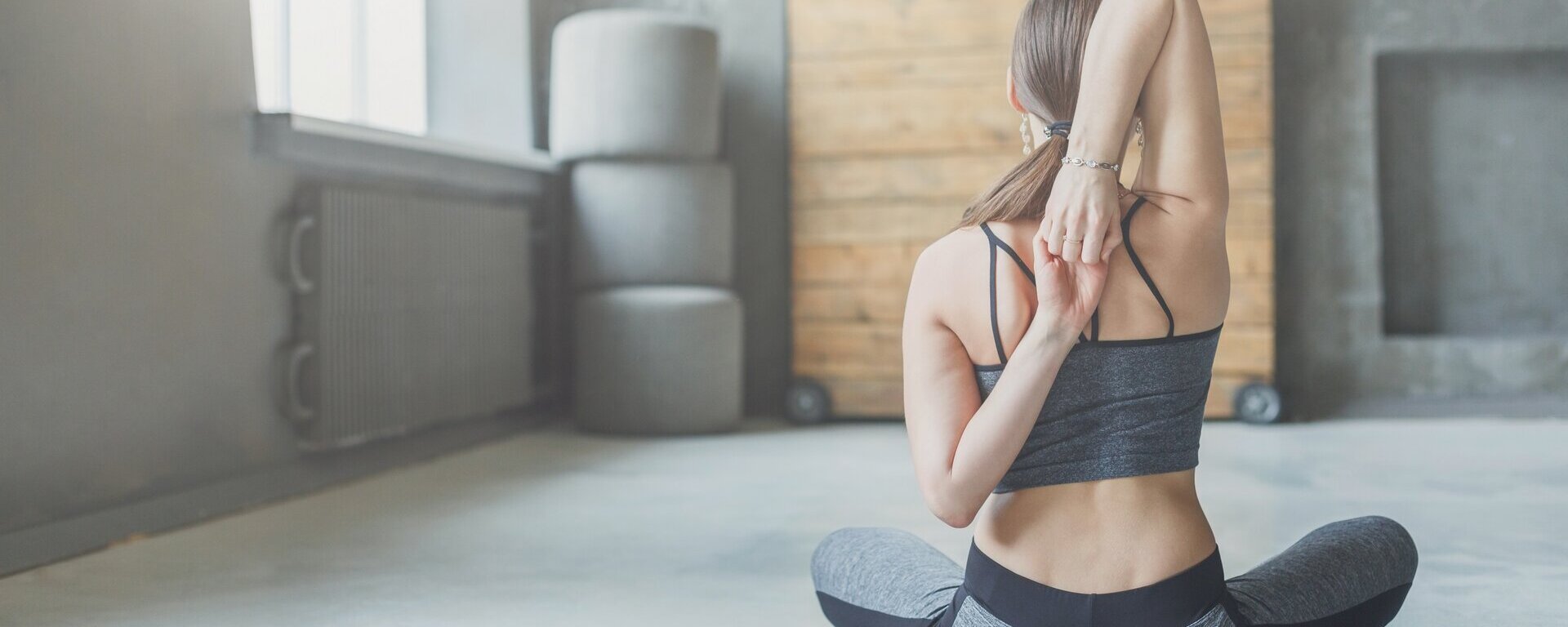 Eine Frau sitzt im Schneidersitz in Sportbekleidung in einem Studio. Man sieht ihren Rücken und sie verschränkt die Arme dahinter.