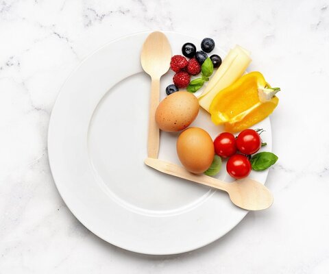 Ein Teller mit zwei Holzlöffeln, was die Optik einer Uhr bildet. Zwischen beiden Löffeln Gemüse, Obst und Eier.