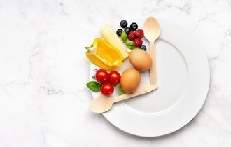 Ein Teller mit zwei Holzlöffeln, was die Optik einer Uhr bildet. Zwischen beiden Löffeln Gemüse, Obst und Eier.